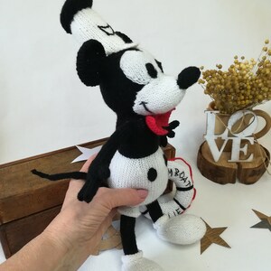 May include: A black and white knitted Mickey Mouse doll wearing a white sailor hat and holding a red and white life preserver. The doll has a red tongue sticking out and black button eyes.