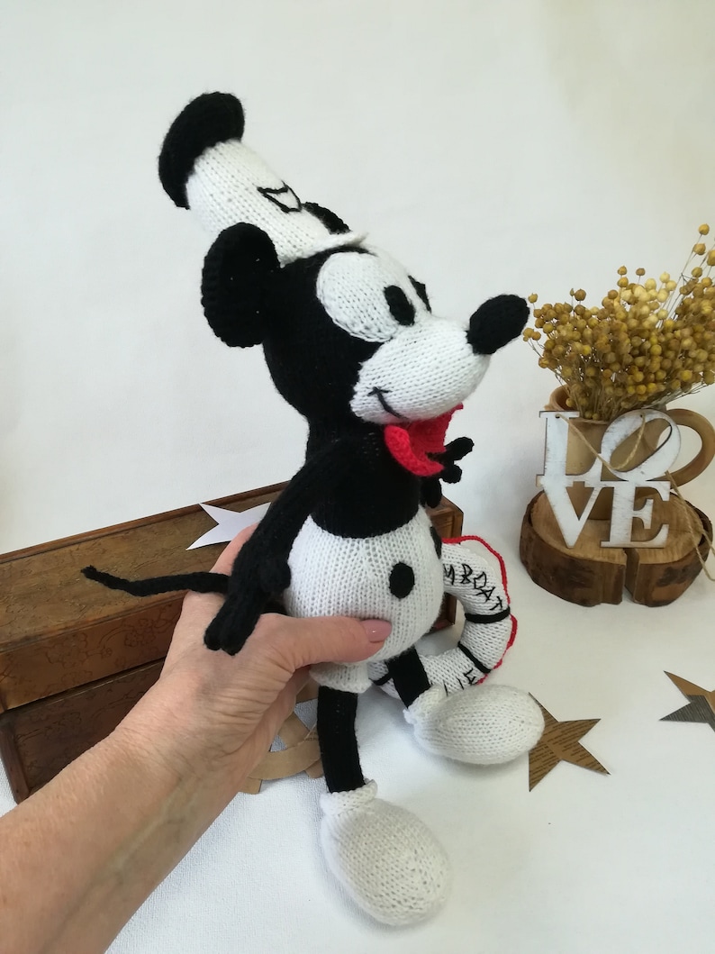 May include: A black and white knitted Mickey Mouse doll wearing a white sailor hat and holding a red and white life preserver. The doll has a red tongue sticking out and black button eyes.