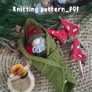 May include: Knitting pattern PDF for a gnome doll wrapped in a green knitted blanket. The gnome is wearing a red and green hat and has a white beard. There is a small knitted snail next to the gnome.