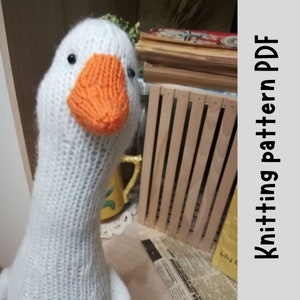 Puede incluir: Juguete de amigurumi de ganso blanco tejido con un pico naranja. El ganso está sentado en una mesa con una caja de madera y un periódico en el fondo. Patrón de tejido PDF.