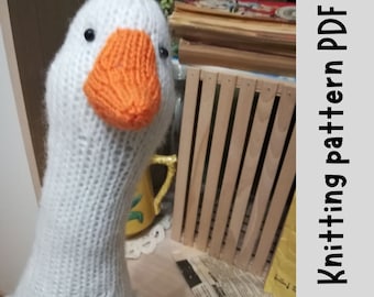 May include: White knitted goose amigurumi toy with an orange beak. The goose is sitting on a table with a wooden crate and a newspaper in the background. Knitting pattern PDF.
