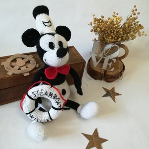 May include: A white and black knitted Mickey Mouse doll wearing a white sailor hat and a red bow tie. The doll is holding a white life preserver with the words "Steamboat Willie" written on it.