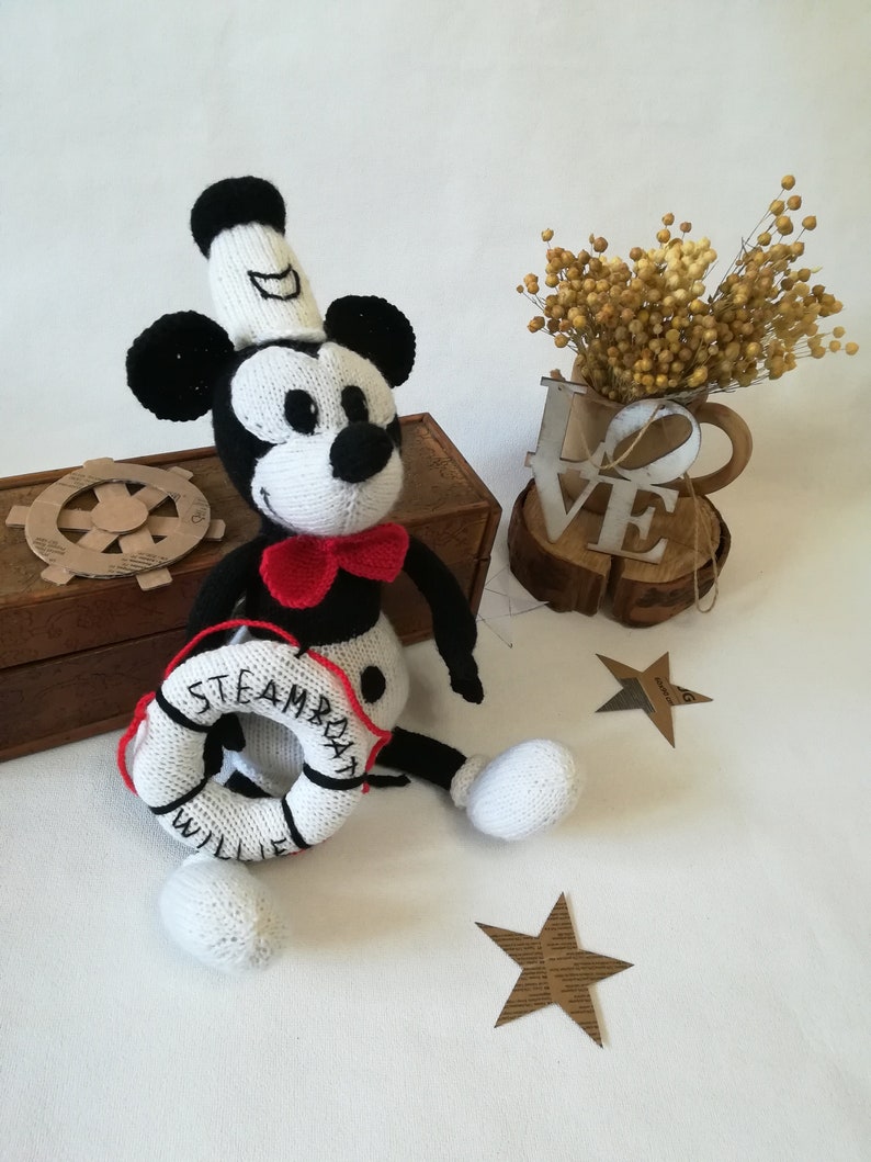 May include: A white and black knitted Mickey Mouse doll wearing a white sailor hat and a red bow tie. The doll is holding a white life preserver with the words "Steamboat Willie" written on it.