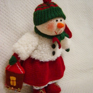 Könnte beinhalten: Eine gestrickte Schneemann-Puppe, die einen grün-roten Hut, einen weißen Pullover, einen roten Rock und rote Stiefel trägt. Der Schneemann hält ein kleines braunes Haus.