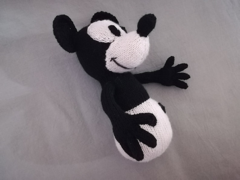 May include: Black and white knitted plush toy resembling a cartoon mouse. The toy has a large, round head with a white face and black eyes. The body is black with white accents.