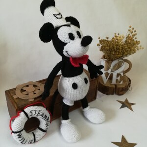May include: A black and white knitted Mickey Mouse doll wearing a white sailor hat and a red bow tie. The doll is sitting on a wooden box with a life preserver that says "Steamboat Willie" in black lettering.