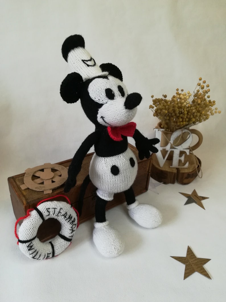 May include: A black and white knitted Mickey Mouse doll wearing a white sailor hat and a red bow tie. The doll is sitting on a wooden box with a life preserver that says "Steamboat Willie" in black lettering.