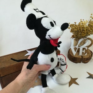 Puede incluir: Un muñeco de Mickey Mouse de punto negro y blanco que lleva un gorro de marinero blanco y sostiene un salvavidas rojo y blanco. El muñeco tiene la lengua roja que sobresale y ojos negros de botón.