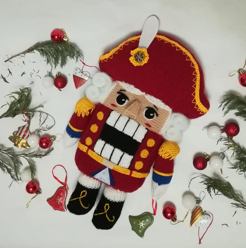 Puede incluir: Un juguete de cascanueces de punto con un gorro rojo y blanco, un uniforme rojo y amarillo y una barba blanca. El juguete est&aacute; rodeado de peque&ntilde;os adornos navide&ntilde;os.