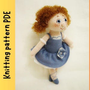 May include: Knitted doll with curly brown hair, blue eyes, and a blue dress with a white lace trim and a flower embellishment. The doll is wearing blue ballet shoes.