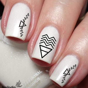 Puede incluir: Esmalte de uñas blanco con diseños geométricos negros y detalles florales. Los diseños incluyen triángulos, zigzags y pequeñas ramitas florales.