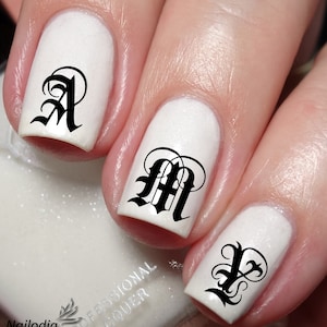 Puede incluir: Esmalte de uñas blanco con calcomanías de letras góticas negras en cada uña. Las letras son A, M e Y.