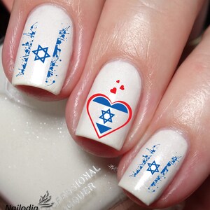 May include: White nail polish with blue and red nail art featuring the Israeli flag. One nail has a heart with the Israeli flag inside. Another nail has the Israeli flag with blue paint splatters.