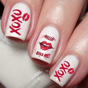 May include: White nail polish with red accents. The nails feature the text "XOXO" and a red lip print. One nail displays the text "HELLO KISS ME!" with a red lip graphic. A romantic and playful nail art design.