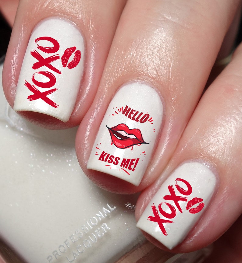 May include: White nail polish with red accents. The nails feature the text "XOXO" and a red lip print. One nail displays the text "HELLO KISS ME!" with a red lip graphic. A romantic and playful nail art design.