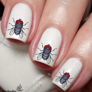 May include: White nail polish with fly nail art. Each nail features a cartoon fly design with red eyes, gray body, and black wings. The image includes the text "Nailodia" and "Proudly Designed and Produced in Australia".