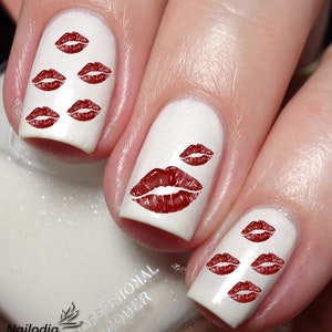May include: White nail polish with red lipstick kiss print nail art. The nails are painted with a glossy white polish and feature multiple red lip prints in various sizes. The design is a close-up shot.