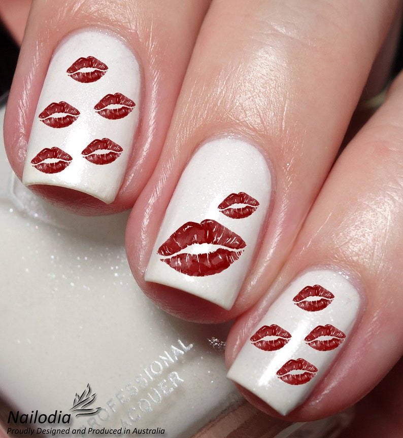 May include: White nail polish with red lipstick kiss print nail art. The nails are painted with a glossy white polish and feature multiple red lip prints in various sizes. The design is a close-up shot.