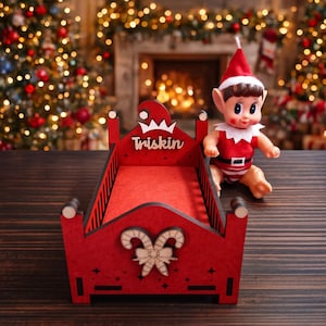 Puede incluir: Una estructura de cama de madera roja con el nombre "Triskin" en la cabecera, decorada con un diseño de bastón de caramelo y lazo. Una muñeca elfo con un atuendo rojo y blanco está sentada junto a la cama. Tema navideño.