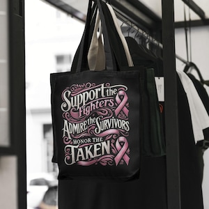 Pu&ograve; includere: Borsa tote nera con la scritta "Support the Fighters, Admire the Survivors, Honor the Taken" in corsivo bianco e rosa. La borsa presenta grafiche a nastro rosa e ha manici beige. La borsa &egrave; appesa a un rack nero con altri vestiti.