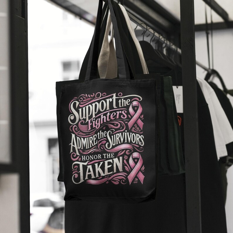 Pu&ograve; includere: Borsa tote nera con la scritta "Support the Fighters, Admire the Survivors, Honor the Taken" in corsivo bianco e rosa. La borsa presenta grafiche a nastro rosa e ha manici beige. La borsa &egrave; appesa a un rack nero con altri vestiti.