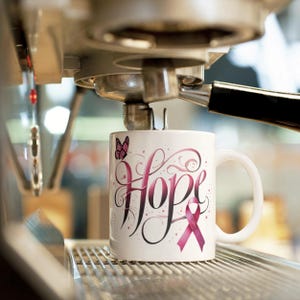Pu&ograve; includere: Tazza in ceramica bianca con la parola "Hope" in corsivo, decorata con volute rosa e nere e un nastro rosa. Una farfalla rosa &egrave; sul lato sinistro della tazza. La tazza &egrave; su una superficie metallica.