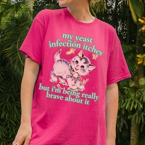 Puede incluir: Una camiseta magenta con el texto "my yeast infection itches but I'm being really brave about it" y un gráfico de gatitos. La persona lleva la camiseta al aire libre, rodeada de vegetación.