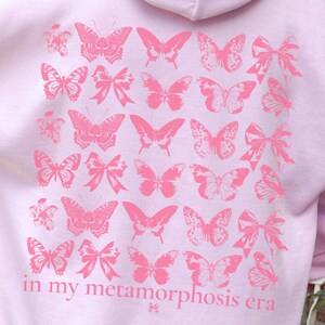 Puede incluir: Una sudadera con capucha rosa claro con capucha y mangas largas. La parte trasera de la sudadera presenta un dise&ntilde;o de mariposas y lazos rosas con el texto "in my metamorphosis era". La sudadera est&aacute; hecha de un material suave.