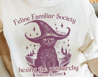 May include: Cream-colored t-shirt with a purple graphic featuring a cat wearing a witch hat, the text "Feline Familiar Society" and "hexing the patriarchy". The design includes stars and the words "Salem Coven".
