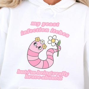 Könnte beinhalten: Weißer Kapuzenpullover mit einer rosa Wurm-Grafik, die eine Blume hält. Der Text auf dem Hoodie lautet "my yeast infection itches but I'm being really brave about it."