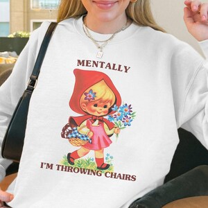 May include: White sweatshirt with a graphic of a vintage-style girl in a red hood holding flowers and a basket. The text "MENTALLY I'M THROWING CHAIRS" is printed above and below the graphic. The sweatshirt is long-sleeved.