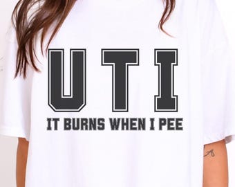 May include: A white short-sleeved t-shirt featuring the black text UTI in a large collegiate font, with the smaller black text IT BURNS WHEN I PEE printed directly underneath. The garment has a crew neckline and a loose, relaxed fit.