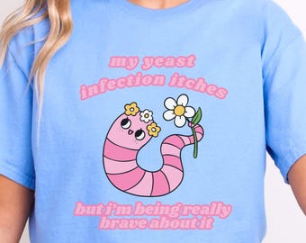 May include: A light blue t-shirt with a pink worm cartoon holding a flower. The text on the shirt reads "my yeast infection itches but i'm being really brave about it".