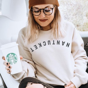 May include: A woman wearing a beige sweatshirt with the text "NANTUCKET" in black letters, a brown beanie, and black jeans. She is holding a white Starbucks cup with the Starbucks logo.