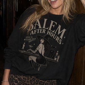 May include: A black long-sleeve sweatshirt featuring a graphic print with the text SALEM AFTER HOURS and OPEN TIL WITCHING HOUR, depicting a witch in a hat sitting at a bar with a black cat, paired with a brown and black leopard print mini skirt.