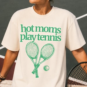 May include: A cream-coloured short-sleeve t-shirt featuring the green text "hot moms play tennis" printed on the chest, accompanied by a graphic illustration of two crossed tennis rackets and a single tennis ball positioned below the text.