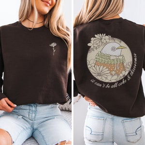 May include: A brown sweatshirt with a small flower design on the front and a larger design on the back. The back design features a bird with a floral pattern and the text "it can't be all coke & threecomes".