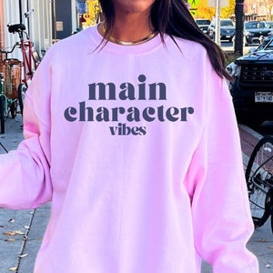 Puede incluir: Una sudadera rosa con el texto "main character vibes" en letras negras.