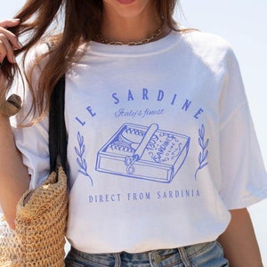 model is wearing comfort colors tee in white with le sardine can and italy's finest direct from sardinia in blue on front