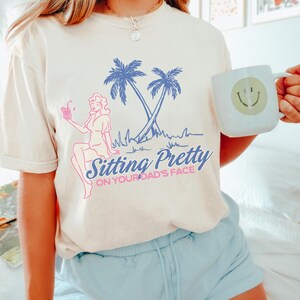May include: Cream-colored t-shirt with a graphic featuring a woman, palm trees, and the text "Sitting Pretty ON YOUR DAD'S FACE" in blue and pink. The design is a retro style. A white mug with a smiley face is held in the background.