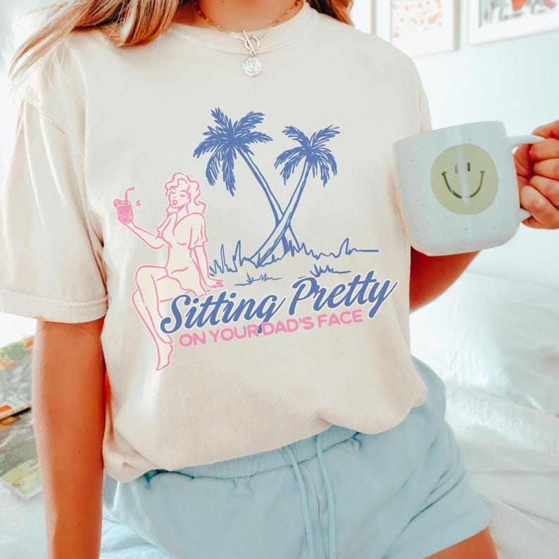 May include: Cream-colored t-shirt with a graphic featuring a woman, palm trees, and the text "Sitting Pretty ON YOUR DAD'S FACE" in blue and pink. The design is a retro style. A white mug with a smiley face is held in the background.