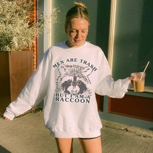 May include: White sweatshirt featuring a raccoon graphic and the navy blue text "MEN ARE TRASH BUT I AM A RACCOON". The raccoon illustration is black and white. A clear plastic cup with a straw is held.