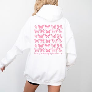 Puede incluir: Sudadera blanca con un dise&ntilde;o de mariposas y lazos rosas en la espalda. El texto "in my metamorphosis era" est&aacute; impreso debajo. La sudadera tiene capucha y mangas largas.