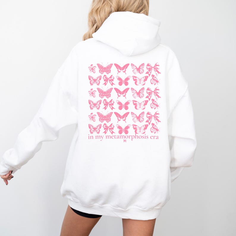 Puede incluir: Sudadera blanca con un dise&ntilde;o de mariposas y lazos rosas en la espalda. El texto "in my metamorphosis era" est&aacute; impreso debajo. La sudadera tiene capucha y mangas largas.