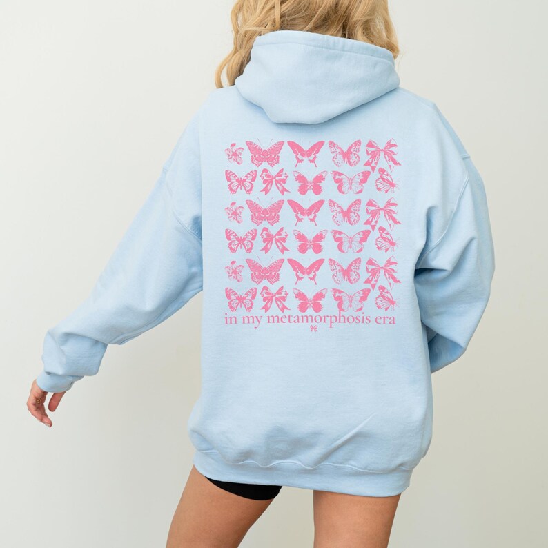 Puede incluir: Sudadera con capucha azul claro con un dise&ntilde;o de mariposas y lazos rosas en la espalda. El texto "in my metamorphosis era" est&aacute; impreso debajo. La sudadera tiene capucha y mangas largas.