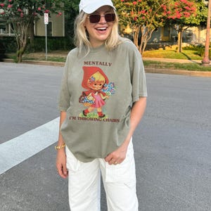 May include: Olive green t-shirt with the text "MENTALLY I'M THROWING CHAIRS" and a graphic of a cartoon girl in a red hood holding flowers. The shirt is paired with white pants and a white cap.