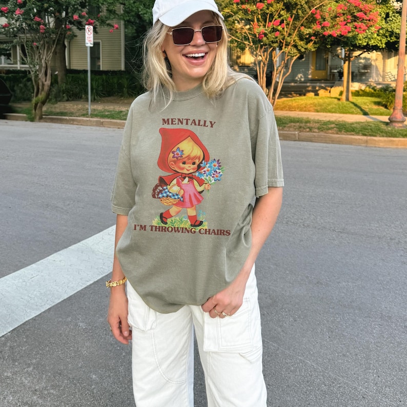 May include: Olive green t-shirt with the text "MENTALLY I'M THROWING CHAIRS" and a graphic of a cartoon girl in a red hood holding flowers. The shirt is paired with white pants and a white cap.