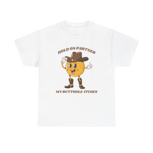 Model is wearing a gildan tee shirt in white with a smiling cowboy giving a thumbs up and it says hold on partner my butthole itches on front in brown