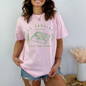 model is wearing comfort colors oversized tee in blossom (light pink) with le sardine can and italy's finest direct from sardinia in green on front