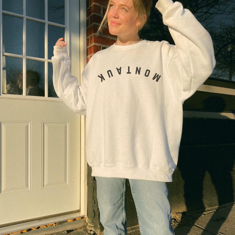 model is wearing gildan oversized crewneck sweatshirt in white with upside down montauk in black across front